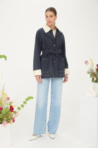 Woman wearing a navy coat with white collar and cuffs, standing against a white background with floral decorations.